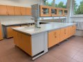 Stół laboratoryjny wyspowy wyposażony w nadstawkę z szafkami przeszklonymi przelotowymi, stanowisko do mycia dwukomorowe, ociekacz kołkowy typu jeż. W tle stoły laboratoryjne przyścienne z szafkami mo